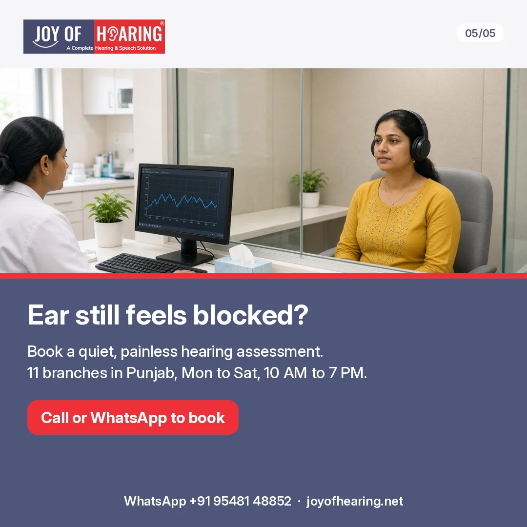 A woman in headphones sits in a hearing test booth while a clinician works at a computer outside the glass.